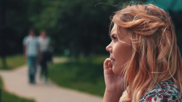 Νεαρή γυναίκα κάθεται στον πάγκο και μιλώντας στο τηλέφωνο στο πάρκο. Ηλιόλουστη μέρα του καλοκαιριού. — Αρχείο Βίντεο