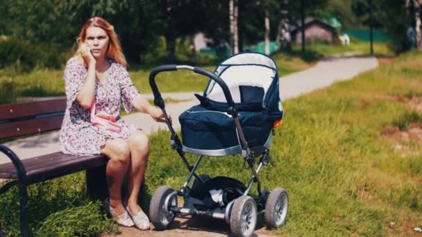 Madre domina el cochecito de bebé y habla por teléfono en el banco en el parque de verano. Soleado. . — Vídeos de Stock