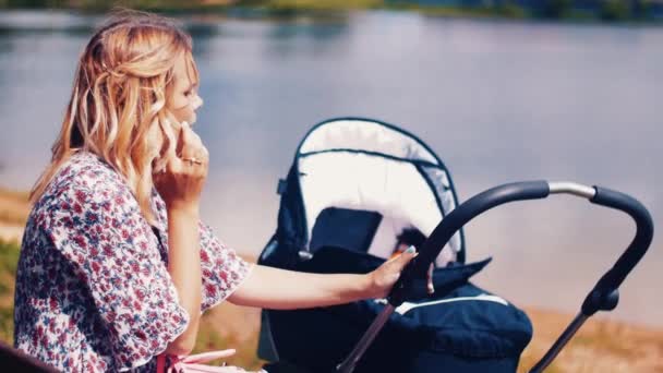 Moeder sway kinderwagen en spreken op telefoon in zomer park Lake. Zonnige. — Stockvideo