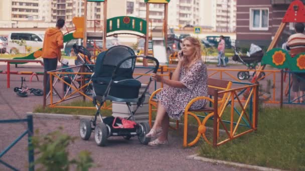 Mutter schwingt Kinderwagen auf Kinderspielplatz und telefoniert. Menschen. — Stockvideo