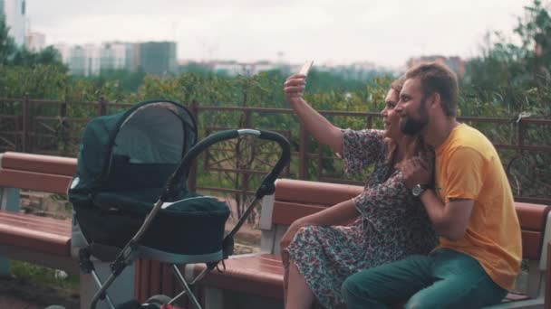 Mother and father with baby carriage on bench. Take selfie on smartphone. Family — Stock Video