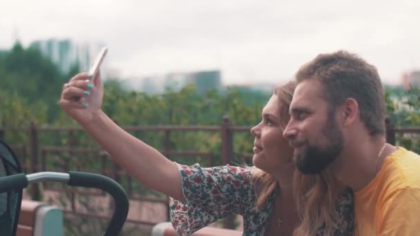 Padres jóvenes con carro de bebé en el banco. Tome selfie en el teléfono inteligente. Familia — Vídeos de Stock