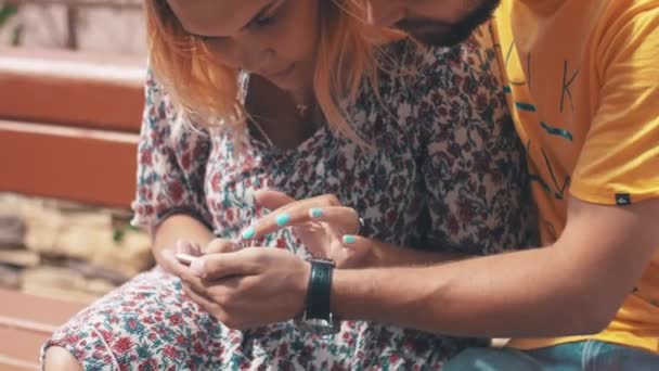 Gelukkig in de liefde paar gebruikend smartphone op Bank. Zomer winderige dag. Zonnige. — Stockvideo