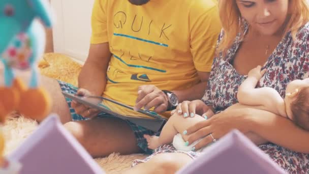 Les jeunes parents jouent à la maison en tablette sur le lit. Maman tient bébé endormi. Jouets . — Video