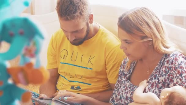 Les jeunes parents à la maison jouent dans le jeu sur tablette. Maman tient bébé endormi. Famille — Video