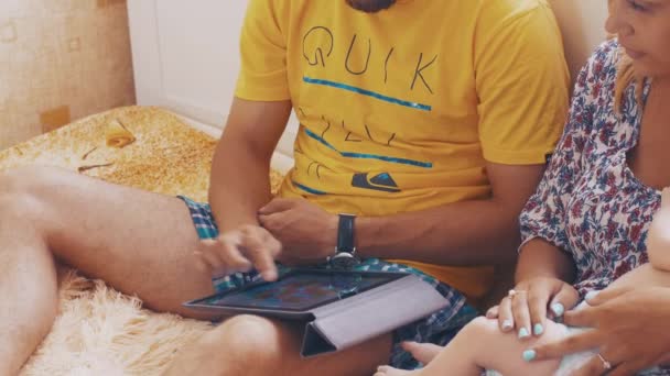 Die Eltern spielen gemeinsam ein Spiel auf dem Tablet. Mutter hält kleines Baby auf Händen — Stockvideo