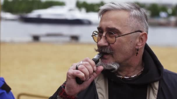 Hombre barbudo retirado fuma cigarrillo electrónico en la playa en primera línea de mar. Vaper. . — Vídeos de Stock