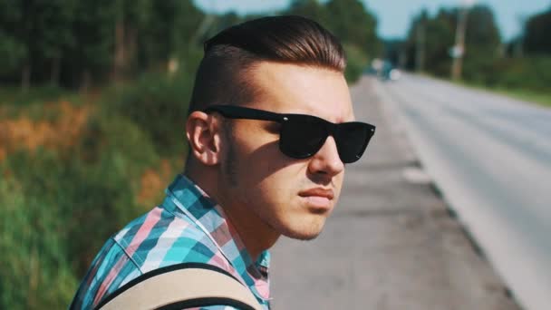 Młody chłopak w okulary autostop w road w letni dzień. Turystycznych. Kciuk w górę. — Wideo stockowe