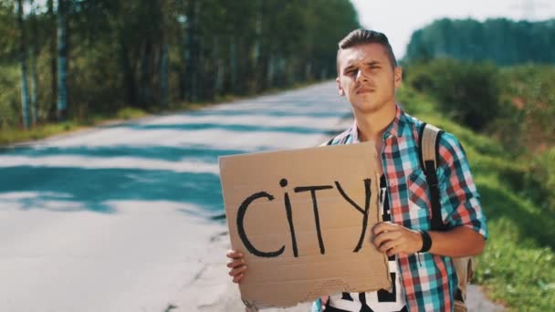Chico sostener placa de cartón con señal de autoestop ciudad en la carretera en el día de verano. Espera. — Vídeos de Stock