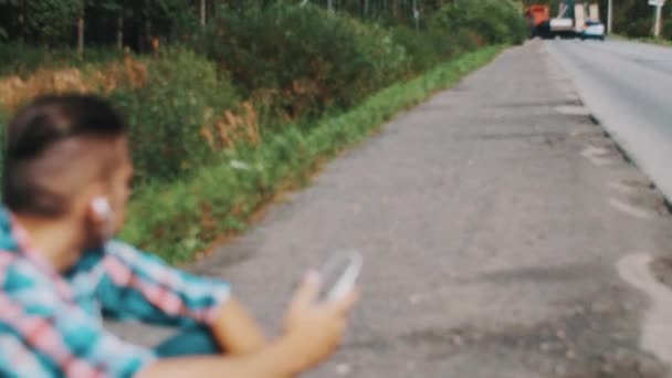 Boy sitting at road listen to music in headphones. Waiting. Cars. Hitchhiking. — Stock Video