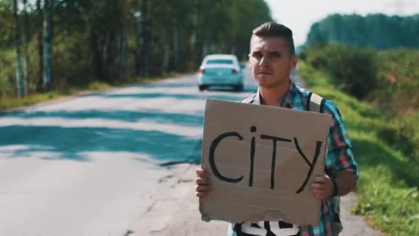 Boy hold placa de papelão com sinal de cidade carona na estrada no verão dia ensolarado . — Vídeo de Stock