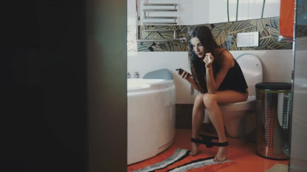 Girl sit on toilet. Black underwear. Tap on phone. Smoke electronic cigarette. — Stock Video