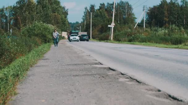 Niño caminando a lo largo de la carretera mantenga placa de cartón con la ciudad signo. Hacer autostop. Automóviles — Vídeos de Stock