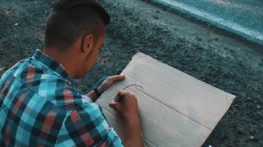 Young boy write word city on cardboard plate sitting at roadside. Hitchhiking