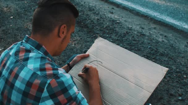 Young boy write word city on cardboard plate sitting at roadside. Hitchhiking — 图库视频影像