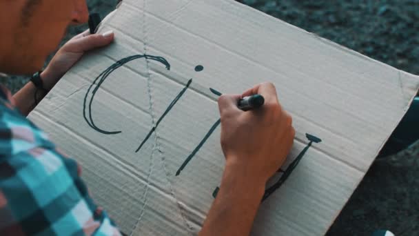 Boy escribir ciudad palabra en placa de cartón por marcador. Sentado en la carretera. Autoestop — Vídeos de Stock