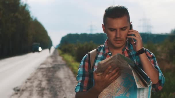 Jongen met rugzak op zoek in de kaart langs de weg. Praat op telefoon. Zonnige dag in de zomer — Stockvideo