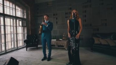 Parlayan elbiseli vokalist saksafoncu ile sahnede performans sergiliyor. Retro tarzı. Caz