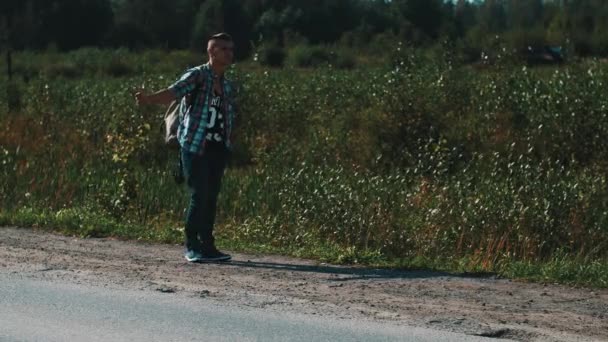 Młody chłopak z plecaka autostopem na poboczu drogi. Podróżnik. Kciuk w górę. Letni dzień — Wideo stockowe