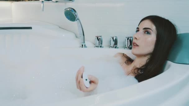 Молодая девушка принимает ванну, полную пены в ванной комнате. Переключение музыки по контроллеру — стоковое видео
