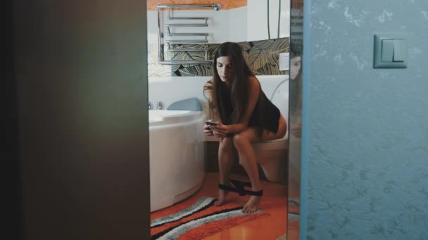 Ragazza bruna sedersi sul water senza toccare biancheria intima in smartphone. Bagno — Video Stock