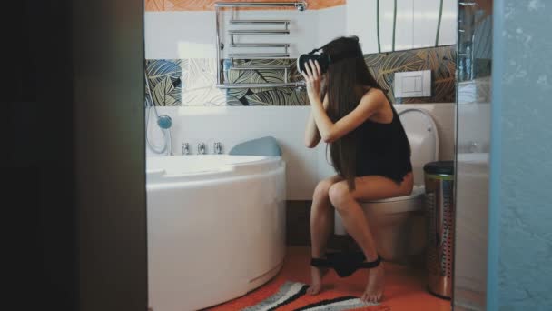 Flickan sitter på toaletten. Titta i virtual reality-glasögon. Underkläder. Spela spelet — Stockvideo