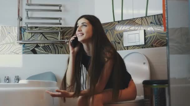 Μελαχρινή κοπέλα, κάθεται στην τουαλέτα συναισθηματικά μιλώντας στο τηλέφωνο. Μπάνιο. Χαμόγελο — Αρχείο Βίντεο