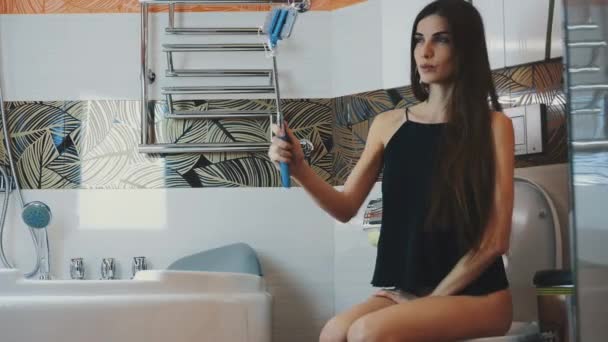 Young girl sitting on toilet in bathroom. Take selfie on blue monopod. Posing. — Αρχείο Βίντεο