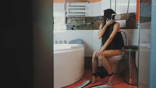 Молодая девушка сидит на унитазе. Смотрю в очки виртуальной реальности. В ванной. Device . — стоковое видео