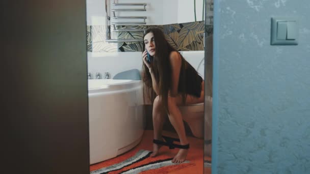 Chica joven sentada en el inodoro sin ropa interior hablando por teléfono. Cuarto de baño. Sonríe — Vídeo de stock