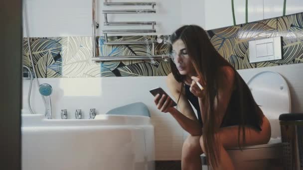 Брюнетка сидит на унитазе со смартфоном. Курение электронных сигарет . — стоковое видео
