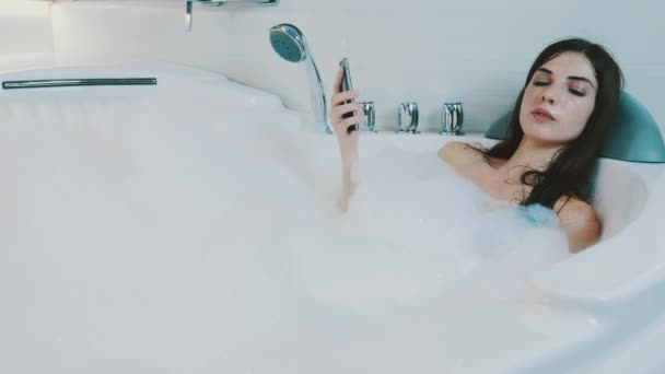 Chica morena disfrutar de un baño lleno de espuma. Toque en el teléfono inteligente. Relájate. Descanso . — Vídeo de stock