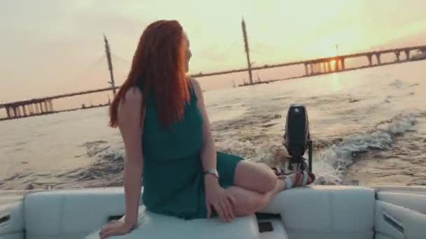 Mladá dívka v tyrkysových šatech sedět na okraji motorového člunu. Západ slunce. Romantický. — Stock video