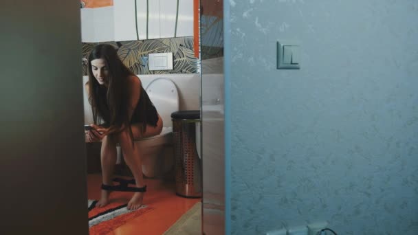 Девушка сидит на унитазе без нижнего белья, постукивая в смартфон. В ванной. Главная страница — стоковое видео