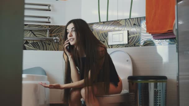 Молодая девушка с длинными темными волосами сидит на унитазе и говорит по телефону. Ванная комната . — стоковое видео