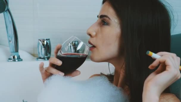 Flicka koppla av i badet full av skum. Röka elektronisk cigarett, dricka rött vin. — Stockvideo
