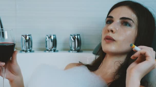 Dívka relaxaci ve vaně plné pěny. Kouření elektronické cigarety, držet červené víno. — Stock video