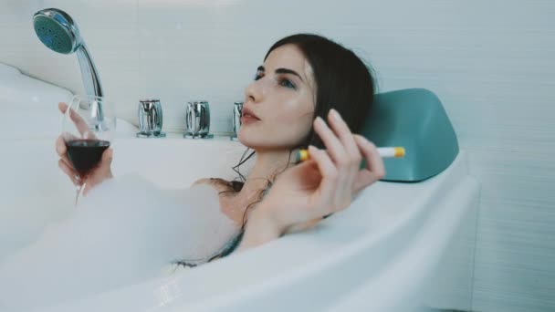 Menina beber vinho tinto no banho cheio de espuma. Fumar cigarro electrónico. Sorria. — Vídeo de Stock