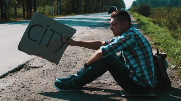 El hombre se sienta en la carretera en el campo con placa de cartón signo de la ciudad. Autoestop . — Vídeo de stock