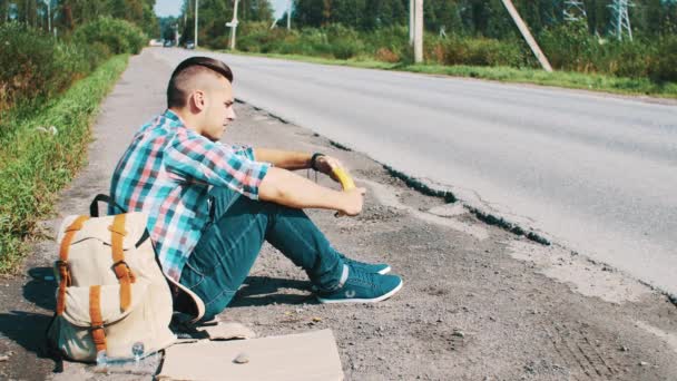 Człowiek siedzieć na drodze w okolicy. Autostop. Czekam na pomoc. Jedzenie banana. — Wideo stockowe