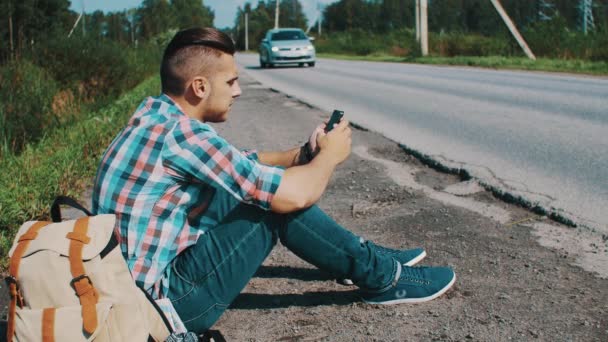 田舎の道で男が座っています。ヒッチハイク。待っています。携帯電話を拾います。バックパック — ストック動画