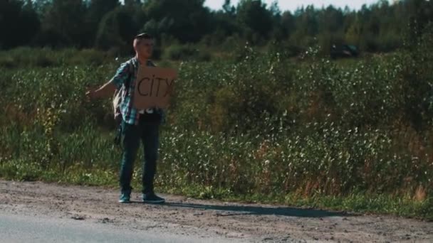 O homem fica na estrada no campo. A pedir boleia. Polegar para cima. Placa de papelão cidade — Vídeo de Stock