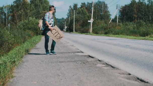 Człowiek spacerem wzdłuż drogi z tektury płyty z znak miasta. Autostop. — Wideo stockowe
