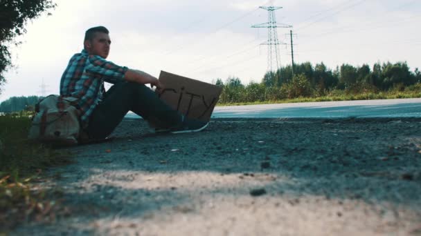 Man sit at road in countryside with cardboard plate sign city. Hitchhiking. Wait — Stock Video