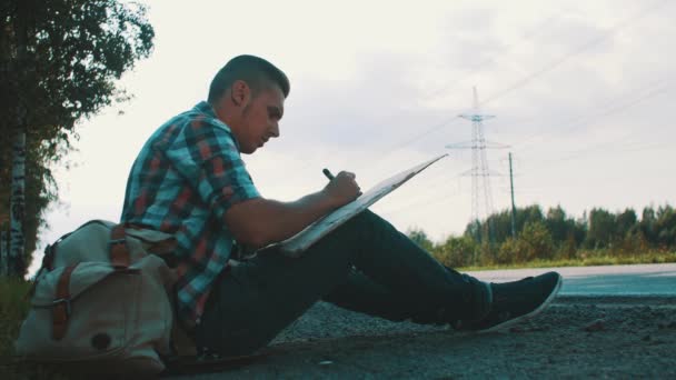 El hombre se sienta en la carretera en el campo escribir en la placa de cartón. Hacer autostop. Esperando. . — Vídeo de stock