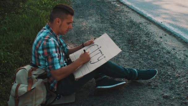 男は、段ボールの板に田舎書き込み単語都市の道路に座っています。ヒッチハイク. — ストック動画