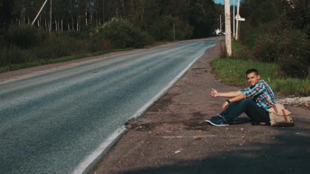 Homem com mochila sentado na estrada no campo. A pedir boleia. Polegar para cima. Viagens — Vídeo de Stock