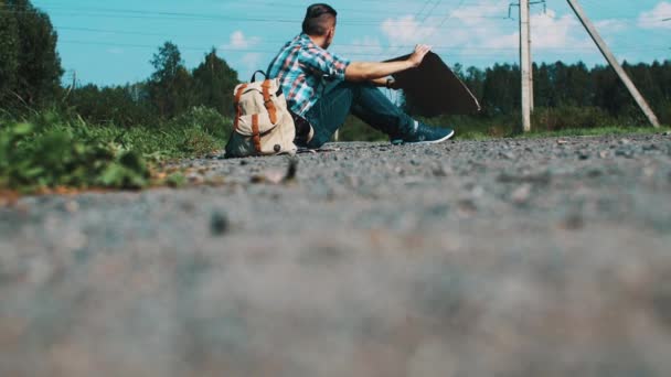 Jovem se senta na estrada no campo com placa de papelão. A pedir boleia. Problema. — Vídeo de Stock