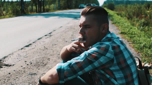 Young man sit at road in countryside. Hitchhiking. Waiting. Smoking cigarette. — Stock Video
