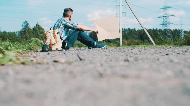 Young man sit hitchhiking at road in countryside hold cardboard plate in hands. — Stock Video